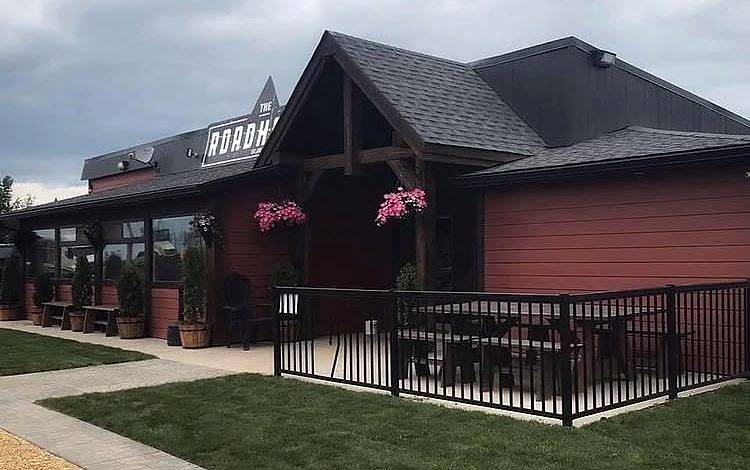 The Roadhouse on a summer day: a red wooden building with the ROADHOUSE sign, a timber-framed entrance with hanging pink flower baskets, and a fenced patio with picnic tables beside a fresh lawn